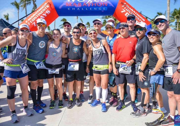 Group of runners with and without physical disabilities pose together beneath a World Marathon Challenge finish arch, wearing race bibs and event apparel in a sunny outdoor setting.