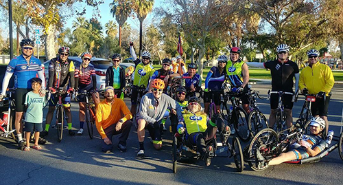 A large group of cyclists, including handcyclists and upright cyclists, gathers outdoors for a group photo before a ride.