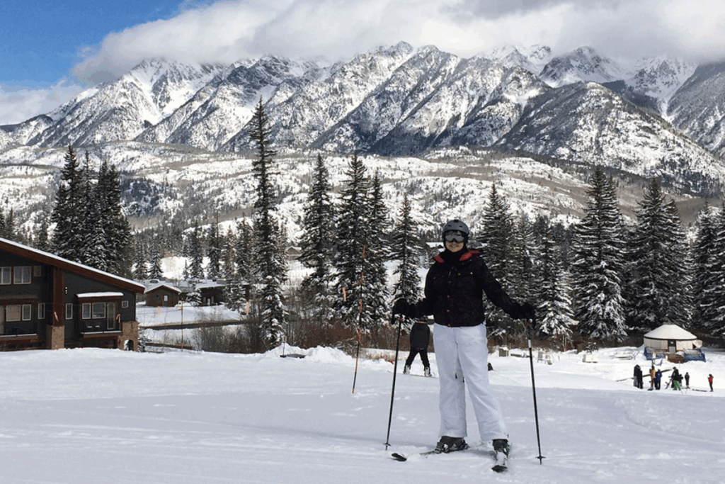 A skier stands on a snowy slope with poles in hand, with snow-covered mountains and pine trees in the background.