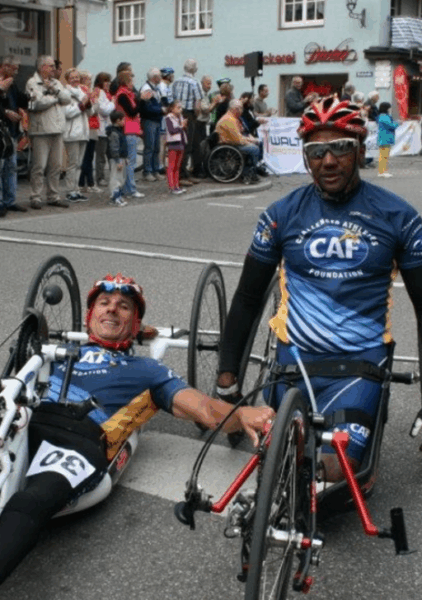 Two handcycle athletes wearing matching CAF race jerseys and helmets pause at a finish-line area during an event. One athlete sits in a white handcycle while the other stands beside a red one, with spectators gathered behind them.