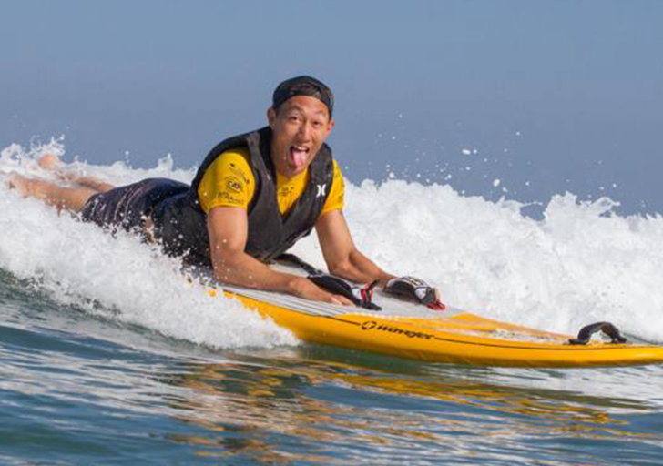 An athlete rides a wave on a yellow adaptive surfboard, smiling with excitement during an ocean surf session.