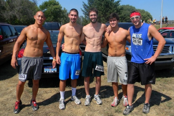 A group of five friends pose together after a race, standing shirtless with race bibs in a parking area.