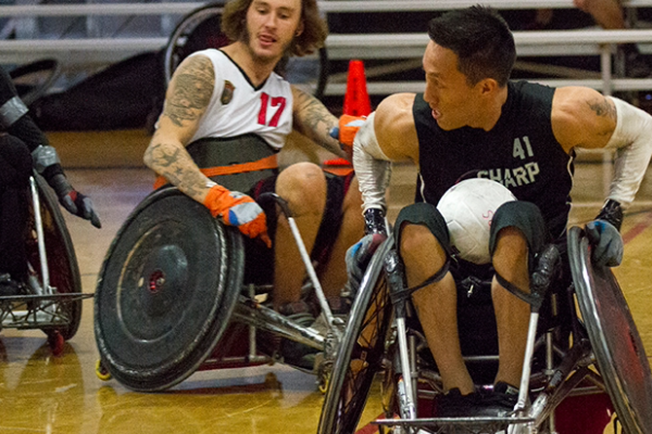 Two athletes compete in a wheelchair rugby match, maneuvering their chairs and fighting for ball control on the court.