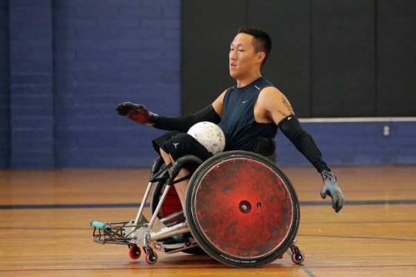 An athlete moves quickly across the court in a wheelchair rugby game, controlling the ball and preparing to pass.