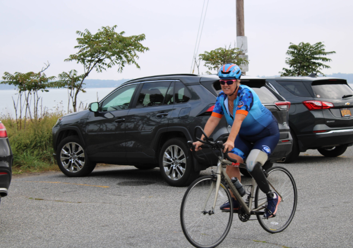 Athlete wearing a blue cycling kit and helmet rides a road bike on a paved road next to parked cars with a body of water in the background