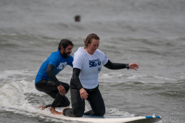 Two individuals surf together on a board in shallow ocean water wearing surf shirts and wetsuits as small waves break around them