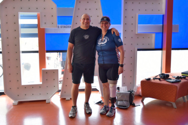 Two individuals stand together indoors in front of large illuminated CAF letters with a bag and camera equipment on the floor nearby