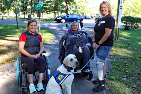 Three individuals gather on a paved path in a park setting with a service dog wearing a vest positioned in the foreground and one individual using a wheelchair