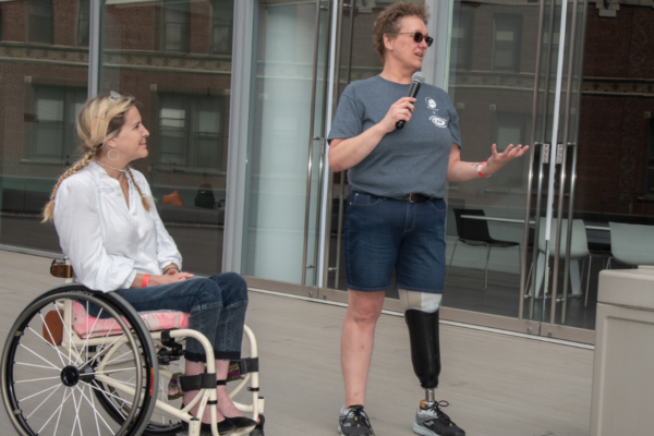 Individual with a prosthetic leg holds a microphone while speaking next to another individual seated in a wheelchair in an outdoor plaza area with glass doors behind them