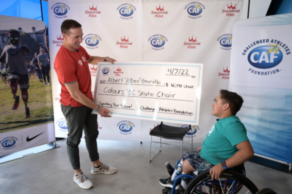 CAF representative presents a large grant check to a youth athlete with a physical disability seated in a wheelchair during a Smoothie King and Challenged Athletes Foundation partnership event.