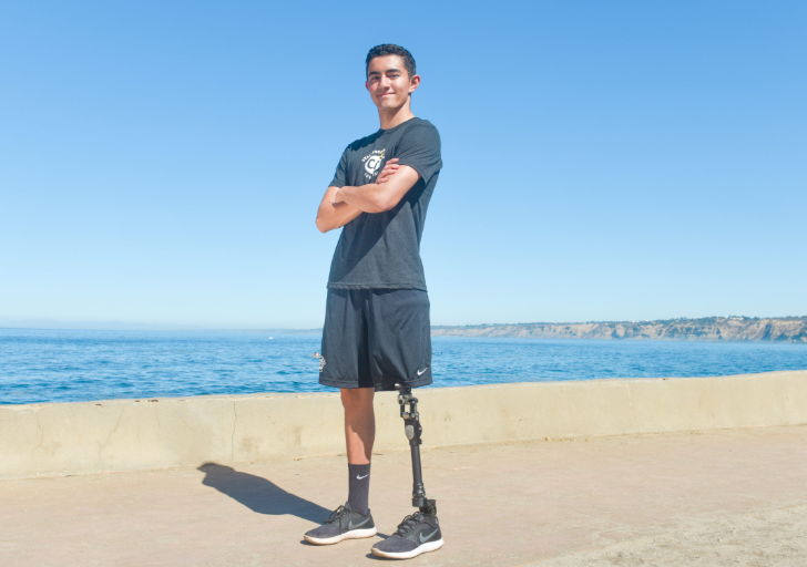 An athlete with a physical disability stands confidently along a seaside walkway, wearing athletic clothing and a prosthetic leg, with the ocean and coastline in the background.