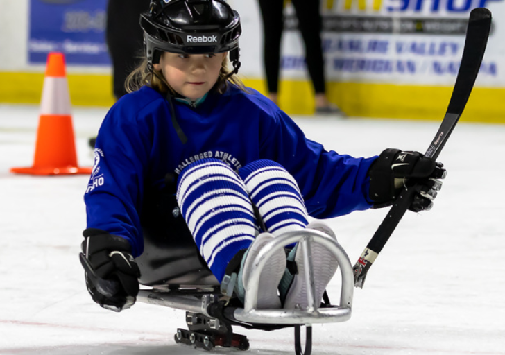 A youth athlete with a physical disability plays sled hockey on an indoor ice rink, wearing a helmet and CAF jersey while holding a hockey stick and gliding across the ice.