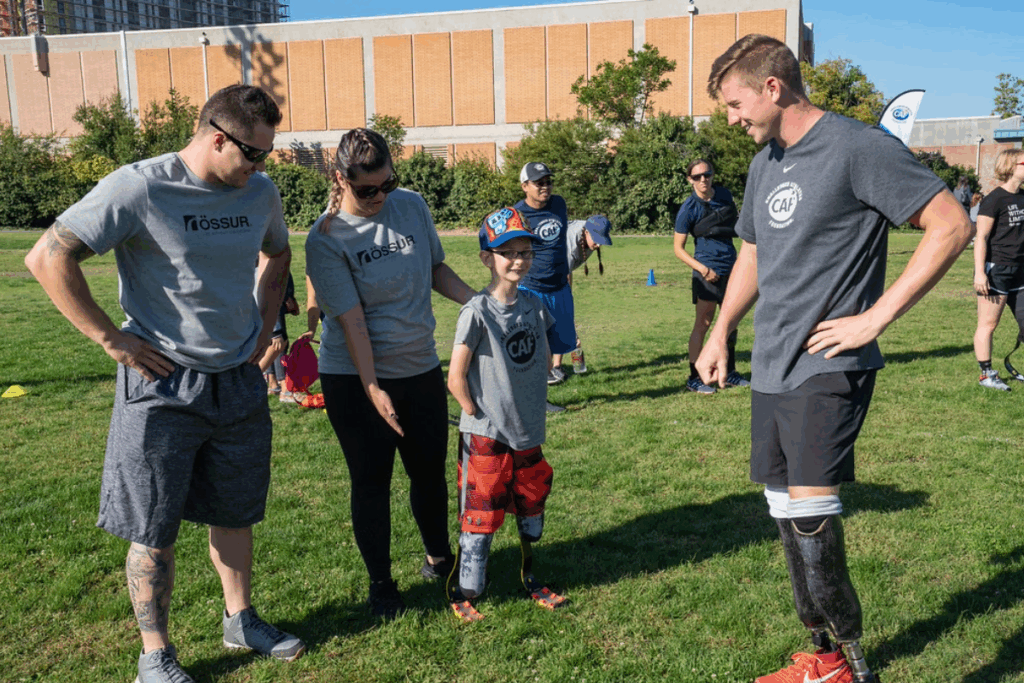 A child with bilateral running blades stands smiling with CAF mentor, Hunter Woodhall, and Össur volunteers on a grassy field.