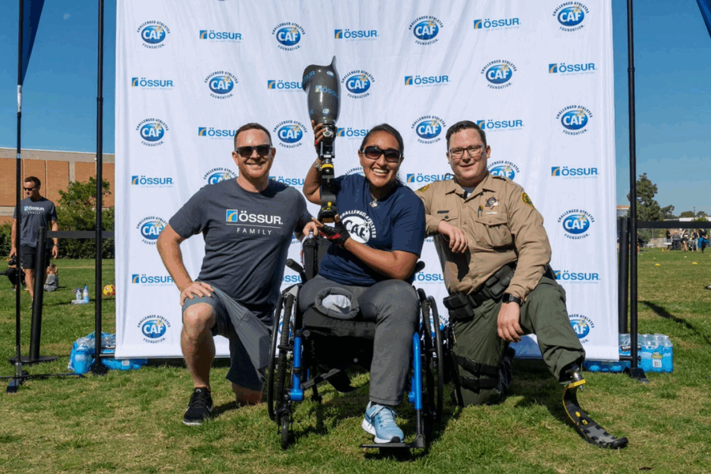 An athlete in a wheelchair raises a running prosthetic with two supporters beside her at an Össur and CAF mobility clinic.