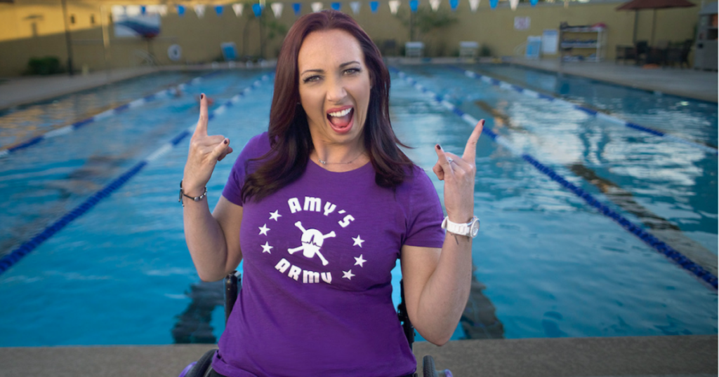An athlete with a physical disability smiles and poses confidently in a wheelchair at the edge of a swimming pool, wearing a purple “Amy’s Army” shirt and raising both hands in a celebratory gesture.