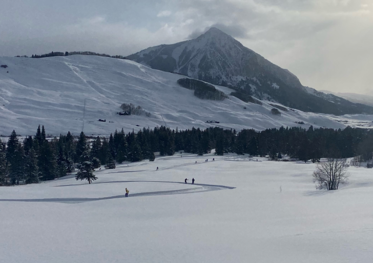 Wide winter landscape showing athletes skiing across a snow-covered valley with winding ski tracks, evergreen trees, and a rugged mountain rising in the background under a cloudy sky.