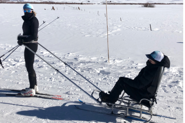 An athlete with a physical disability skis while guiding a sit-ski athlete across a snowy field, using ski poles and a tether system during a winter adaptive sport session.