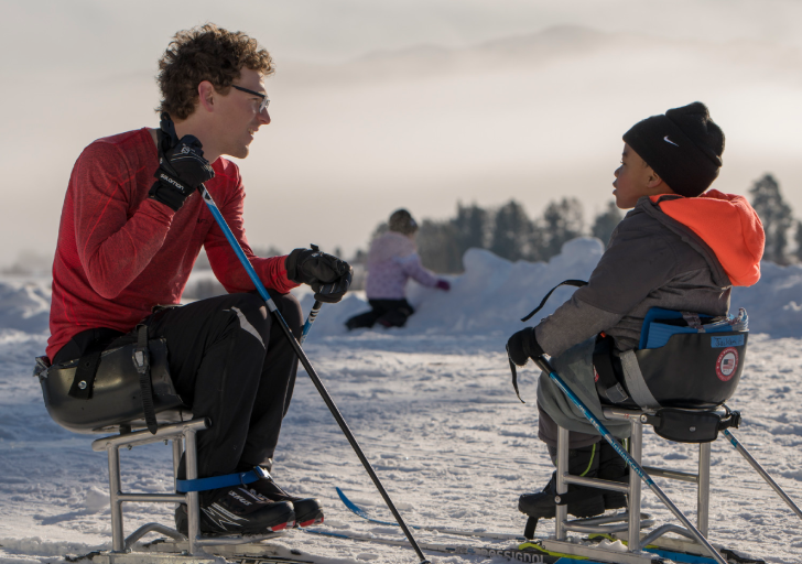 An instructor and a young athlete with a physical disability sit in Nordic sit-skis facing one another on a snowy field, talking and holding ski poles during a winter sports clinic.
