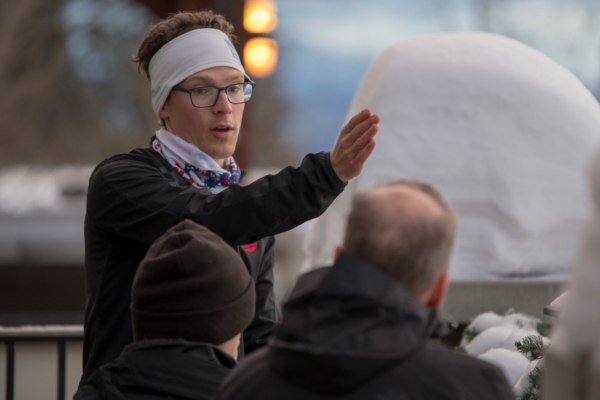 An instructor gestures while giving guidance to participants during a winter sports session, standing near snow-covered equipment and bundled attendees.