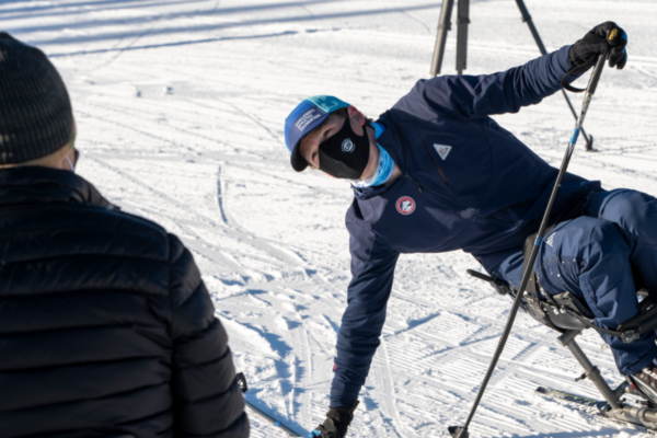 An instructor gestures while giving guidance to participants during a winter sports session, standing near snow-covered equipment and bundled attendees.