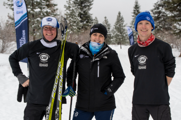 Three CAF-Idaho community members stand smiling on a snowy Nordic trail, holding skis and wearing CAF winter apparel with evergreen trees behind them.