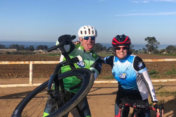 Two cyclists with physical disabilities smile and pose with their bikes during a rest stop, with farmland and the ocean visible in the background.