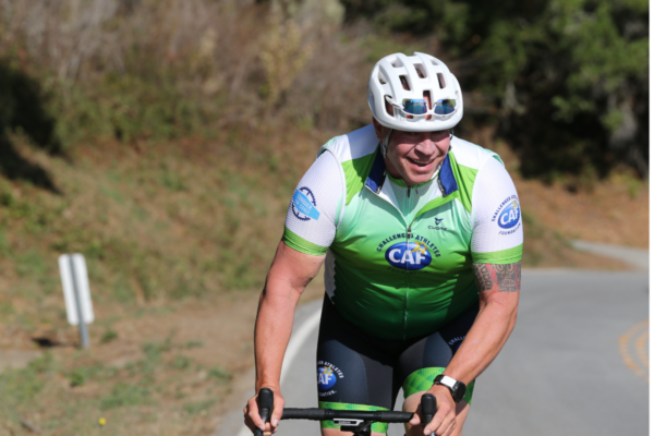 An athlete with a physical disability wearing a CAF cycling jersey rides uphill on a paved road, smiling while pushing through a climb.