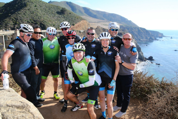 A group of CAF cyclists stand together along a coastal trail, celebrating teamwork and community with ocean cliffs and blue water behind them.