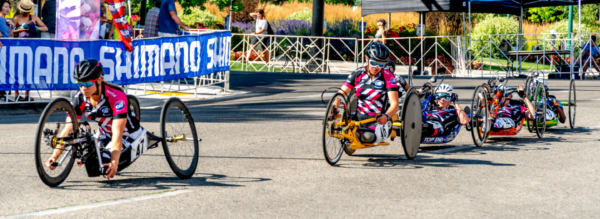 Athletes with physical disabilities compete in a handcycling race, riding low-profile racing handcycles in CAF jerseys as they round a turn on a closed city course.