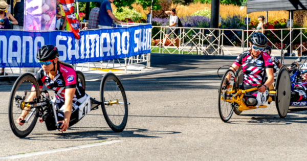 Athletes with physical disabilities compete in a handcycling race, riding low-profile racing handcycles in CAF jerseys as they round a turn on a closed city course.