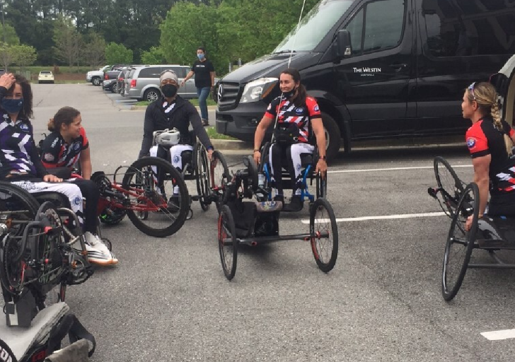 Athletes with physical disabilities gather in a parking lot with handcycles, wearing CAF cycling kits and preparing to ride together.