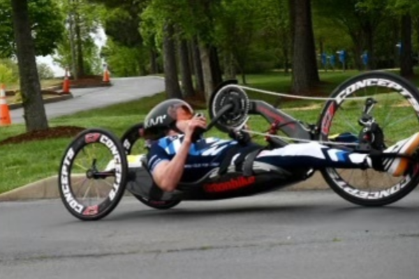 Athletes with physical disabilities pose in wheelchairs holding medals after a paracycling event, wearing CAF cycling kits.