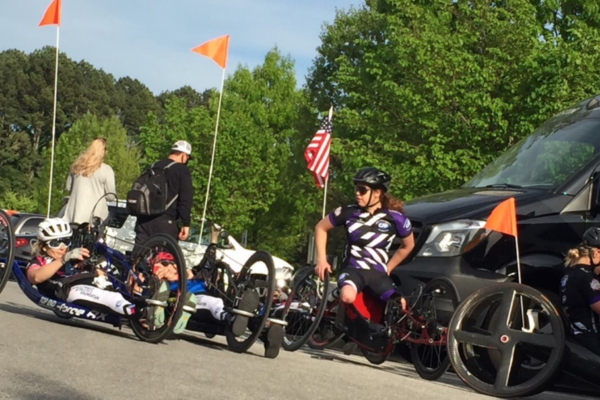 Athletes with physical disabilities line up in handcycles near support vehicles, preparing to begin a paracycling ride.