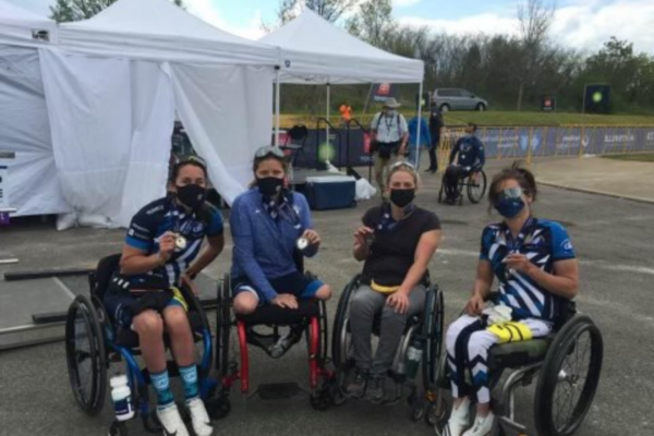 Athletes with physical disabilities pause in handcycles beside a vehicle, adjusting equipment and resting between rides.