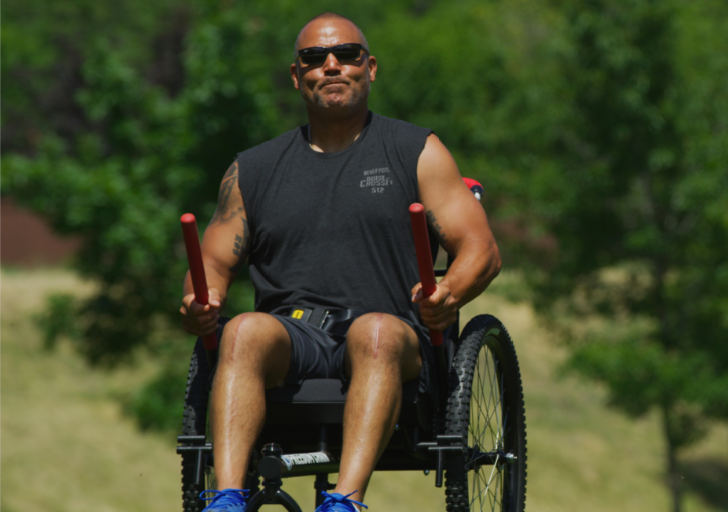 An athlete with a physical disability propels a GRIT Freedom Chair forward using hand grips on a grassy outdoor path.