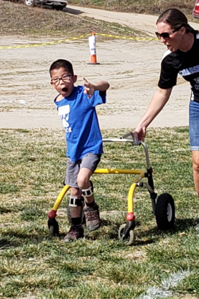 A young athlete with a physical disability practices walking with a mobility walker while supported by an adult on a grassy field.