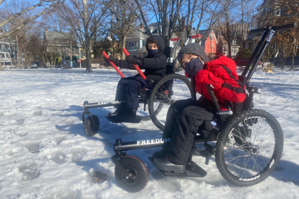 Two young athletes with physical disabilities use GRIT Junior mobility chairs in the snow, bundled in winter clothing at a neighborhood park.