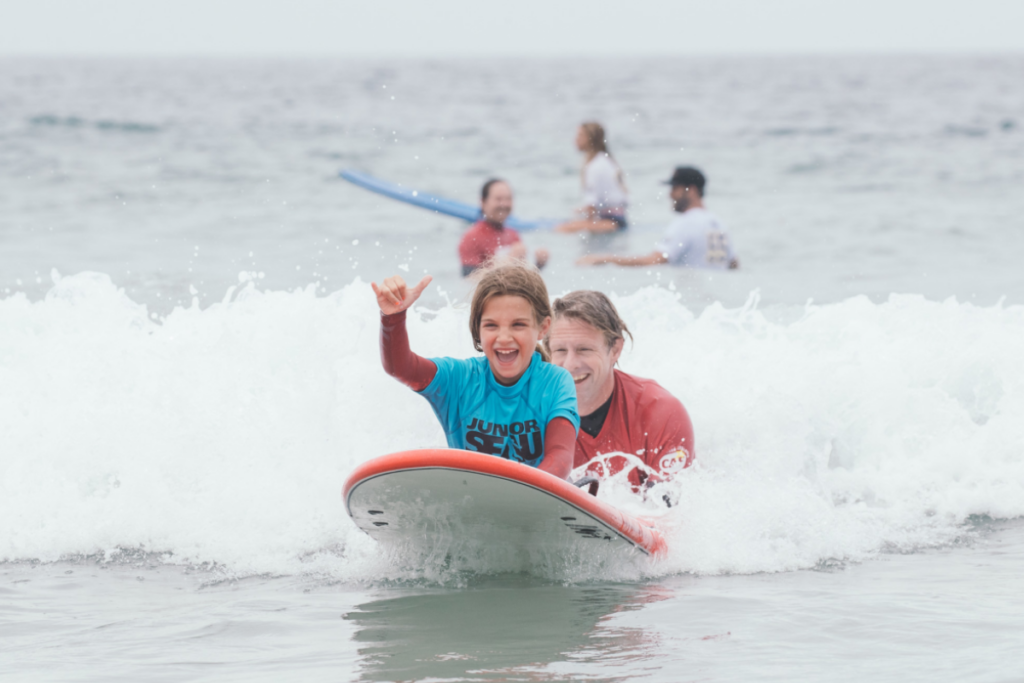 A smiling young athlete with a physical disability rides a surfboard with an instructor, lifting a hand as a wave breaks behind them.