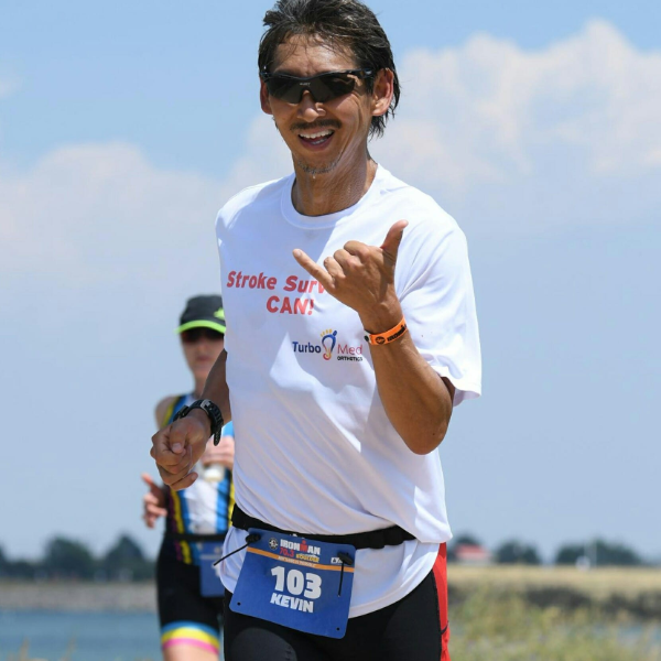 An athlete with a physical disability runs along a lakeside course wearing a race bib during an IRONMAN event.