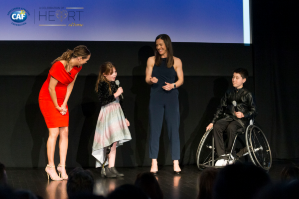Two CAF athletes with physical disabilities and two presenters stand on stage at Celebration of Heart, sharing a lighthearted moment during a live program.