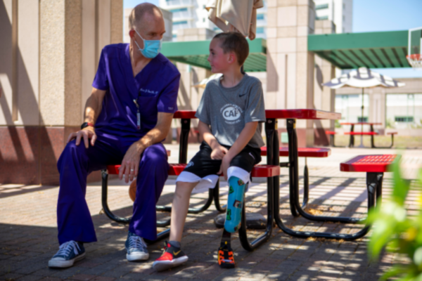 A CAF youth athlete with a physical disability sits on a bench talking with a medical professional, wearing a prosthetic leg and CAF shirt in an outdoor courtyard.