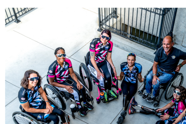 A group of CAF athletes with physical disabilities wearing Team CAF cycling jerseys gather together in wheelchairs, smiling and looking up toward the camera.