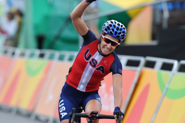 An athlete raises an arm in celebration while riding a road bike in a USA cycling uniform, crossing the finish area during a national championship race.