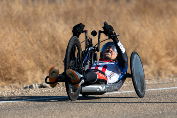 An athlete with a physical disability powers a handcycle down a road, lifting one arm in celebration while competing at the USA Para-Cycling National Championships.