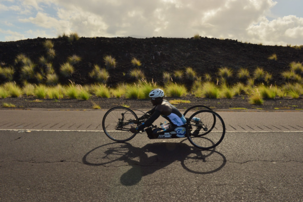 A handcyclist with a physical disability rides along a wide open road surrounded by rugged landscape, competing in a national para-cycling event.