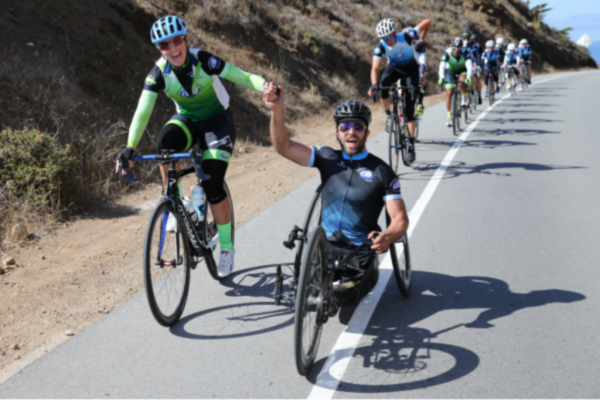 A cyclist and a handcyclist with physical disabilities ride together and clasp hands while a group of cyclists follows behind, highlighting inclusion and camaraderie on the road.