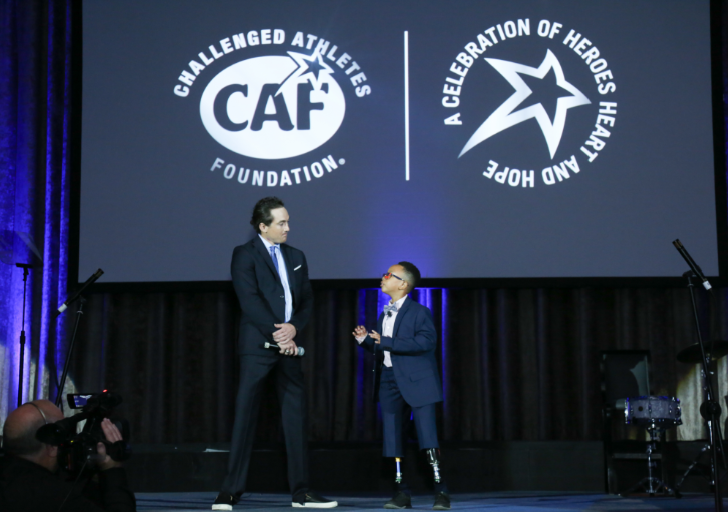 A CAF athlete with a physical disability stands on stage beside a host during Celebration of Heroes, speaking beneath a large screen displaying the Challenged Athletes Foundation and Heart and Hope event logos.
