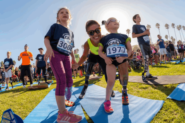 A young girl and a child with a prosthetic leg stand on yoga mats with a smiling adult athlete with prosthetic legs crouching between them, holding hands, as a group of kids and athletes gather behind them at a CAF event.