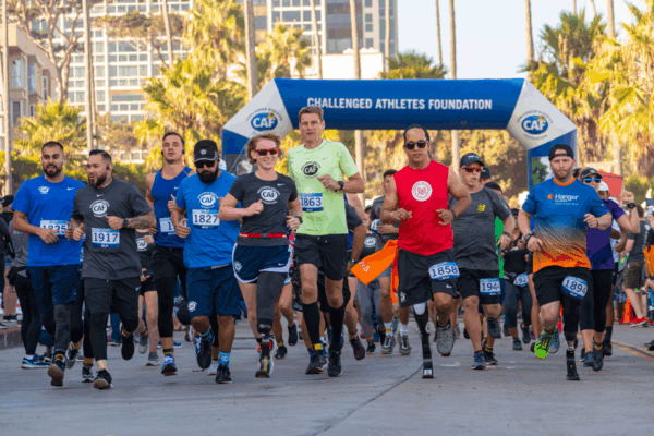 A large group of athletes, including several with running prosthetics, run together from the starting line under a Challenged Athletes Foundation archway at the San Diego Triathlon Challenge.