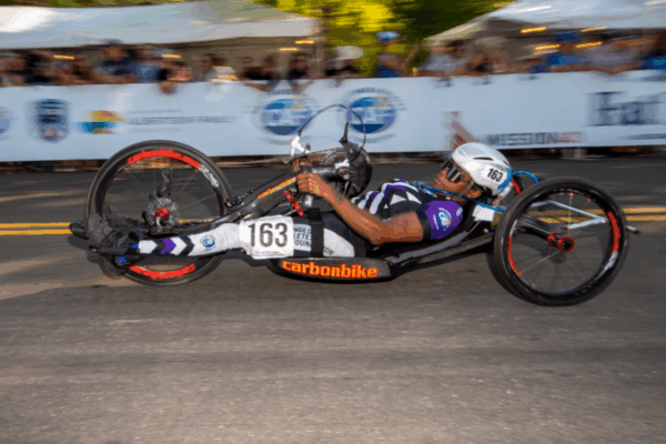 A male athlete with a physical disability races in a black and purple handcycle with spectators cheering behind the barricades.